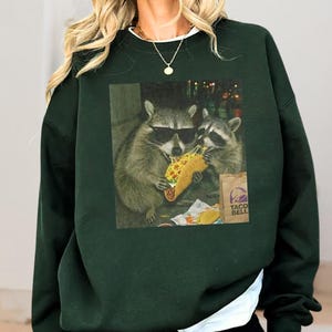 A person wearing a green sweatshirt with a graphic of two raccoons eating tacos.