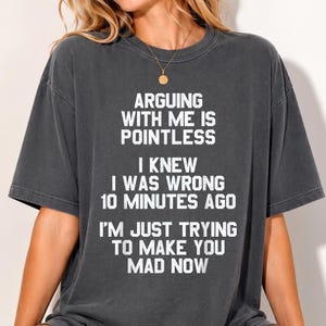 May include: A charcoal gray short-sleeve t-shirt featuring white distressed text that reads: ARGUING WITH ME IS POINTLESS. I KNEW I WAS WRONG 10 MINUTES AGO. I'M JUST TRYING TO MAKE YOU MAD NOW. The shirt has a crew neckline and a relaxed fit.