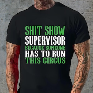 May include: Black t-shirt with the text "SHIT SHOW SUPERVISOR BECAUSE SOMEONE HAS TO RUN THIS CIRCUS" in green and white. The shirt is short-sleeved and made of a soft material.