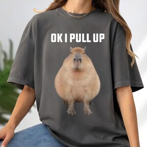 May include: Dark gray t-shirt featuring a large capybara illustration. The text "OK I PULL UP" is printed in white above the capybara. The capybara is light brown with a dark gray nose and feet.