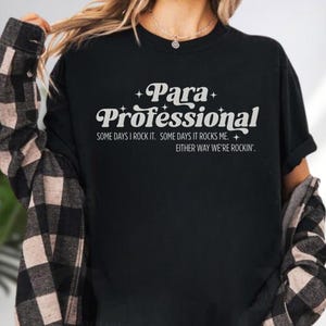 A person wearing a black t-shirt with white text that says "Para Professional" and "Some days I rock it. Some days it rocks me. Either way we're rockin'."