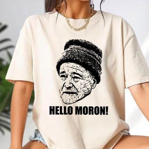 May include: Cream-colored t-shirt featuring a black and white graphic of a man wearing a beanie. The text "HELLO MORON!" is printed in bold, black letters below the image. The shirt has short sleeves and a crew neck.