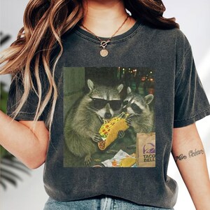 A person wearing a gray t-shirt with a graphic of two raccoons eating a taco.