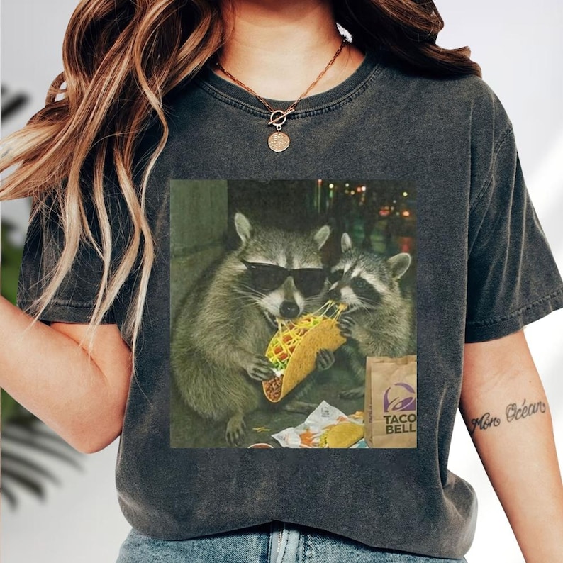 A person wearing a gray t-shirt with a graphic of two raccoons eating a taco.