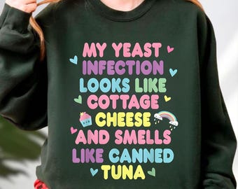 May include: Dark green sweatshirt with the text "MY YEAST INFECTION LOOKS LIKE COTTAGE CHEESE AND SMELLS LIKE CANNED TUNA" in colorful bubble letters. The text is decorated with hearts and a rainbow.