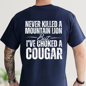 May include: Navy blue t-shirt with white text that reads "NEVER KILLED A MOUNTAIN LION But I'VE CHOKED A COUGAR". The text is centered on the back of the shirt. The shirt is made of a soft material.