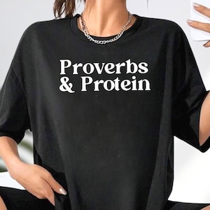 May include: Black t-shirt with the white text "Proverbs & Protein". The shirt has short sleeves and a crew neck. A silver chain necklace is visible at the neckline.