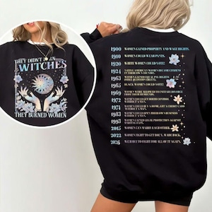 May include: A black sweatshirt featuring a graphic on the back with a timeline of historical dates and text detailing women's rights milestones. The front graphic displays two hands holding a celestial sun and moon design with the text THEY DIDN'T BURN WITCHES THEY BURNED WOMEN.