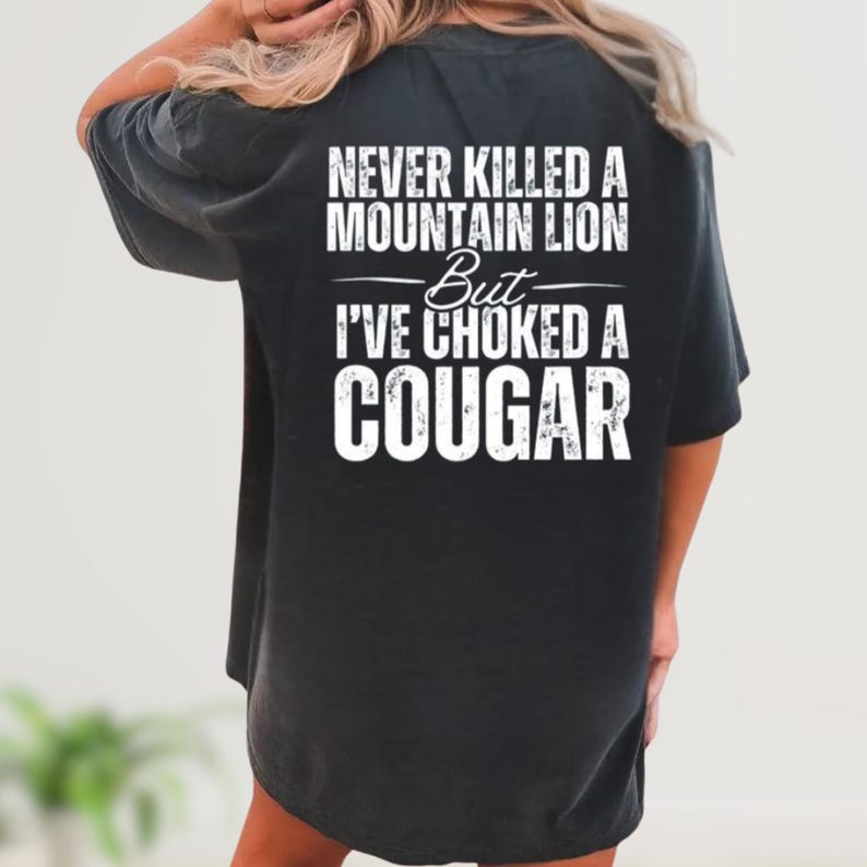 May include: Black t-shirt with white text that reads "NEVER KILLED A MOUNTAIN LION But I'VE CHOKED A COUGAR". The shirt is a relaxed fit.