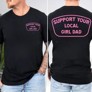 May include: Black t-shirt with the text "SUPPORT YOUR LOCAL GIRL DAD" in pink, displayed on the front and back. The text is inside a pink rounded rectangle. The shirt is a crew neck and short-sleeved.