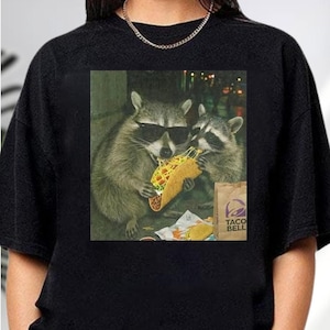 A person wearing a black t-shirt with a graphic of two raccoons eating a taco.