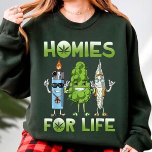 A person wearing a green sweatshirt with a graphic design featuring three cartoon-like characters representing marijuana and the word "HOMIES FOR LIFE".