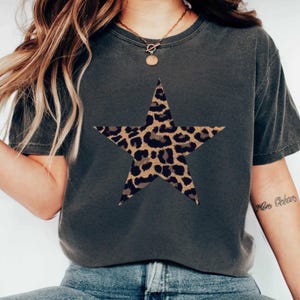 May include: Dark gray t-shirt featuring a large star with a leopard print design. The shirt has short sleeves and a crew neck. The star is centered on the front of the shirt. The shirt is made of a soft, comfortable material.