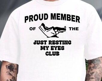 May include: White t-shirt with a black graphic and text. The graphic depicts a person reclining in a chair. The text reads "PROUD MEMBER OF THE JUST RESTING MY EYES CLUB."