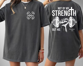 May include: Dark gray t-shirt with white text and graphics. The front features a dumbbell design and the text "Philippians 4:13." The back displays "NOT BY MY STRENGTH BUT HIS" with a cross and dumbbell graphic. A casual, comfortable style.
