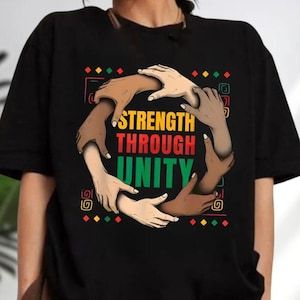 May include: Black t-shirt featuring a graphic with the words "STRENGTH THROUGH UNITY" in yellow, red, and green. The design includes a circle of hands with varying skin tones, symbolizing diversity and togetherness. The shirt has a crew neck.