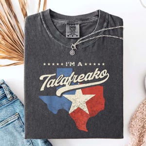 May include: A charcoal gray short-sleeve t-shirt featuring a graphic of the state of Texas in blue, white, and red with a large white star. The text "I'M A Talafreako" is printed above the state outline in a white, stylized, script-like font.