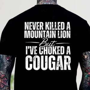 May include: Black t-shirt with white text. The text reads "NEVER KILLED A MOUNTAIN LION But I'VE CHOKED A COUGAR". The shirt has a crew neck and short sleeves. The text is in a bold, sans-serif font.