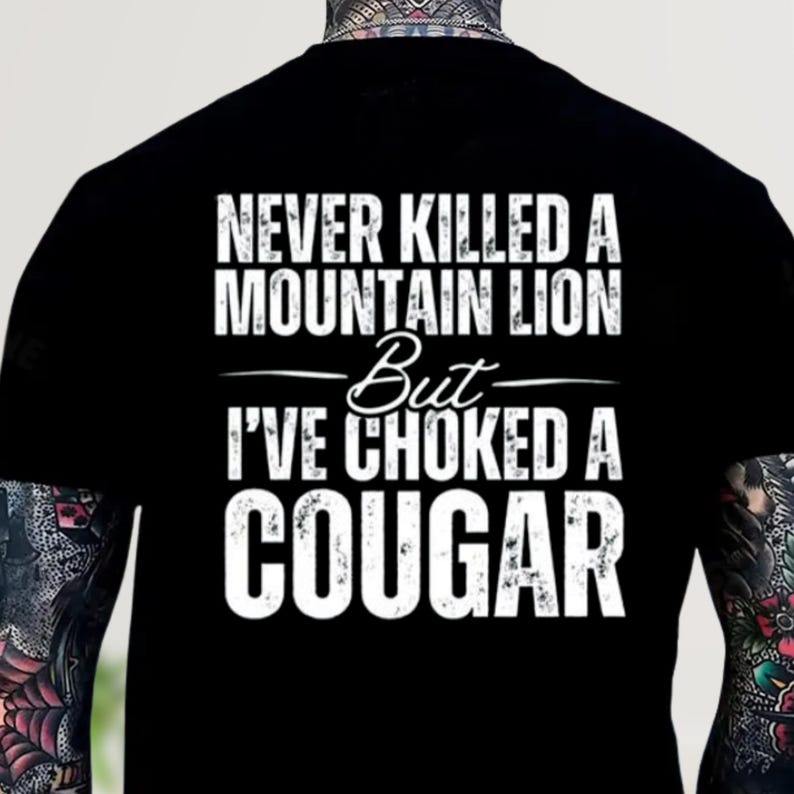 May include: Black t-shirt with white text. The text reads "NEVER KILLED A MOUNTAIN LION But I'VE CHOKED A COUGAR". The shirt has a crew neck and short sleeves. The text is in a bold, sans-serif font.