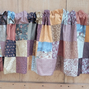 May include: Patchwork valance with a variety of fabrics in shades of brown, purple, and gold. The valance is made of squares of fabric sewn together, creating a rustic, country style. The valance is hanging on a wooden rod.