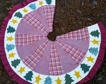 May include: A circular Christmas tree skirt with a burgundy ruffle edge. The skirt features a plaid pattern in red and white, with a border of white fabric adorned with appliqued green trees and yellow stars.