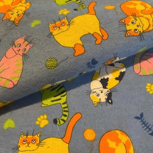 May include: A blue fabric featuring a repeating pattern of cartoon cats in various colors including orange, yellow, pink, and calico. The design includes small yellow paw prints, green yarn balls, and blue fish skeletons scattered across the surface.