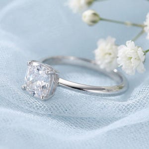 May include: A silver-toned ring featuring a single cushion-cut clear gemstone set in a four-prong setting. The band is smooth and polished, showcasing a classic design with a brilliant, faceted centre stone that reflects light clearly.