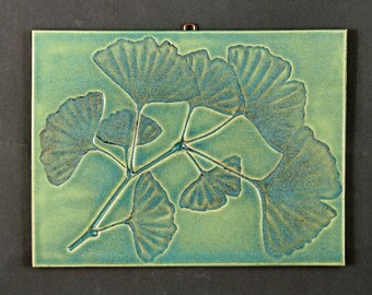 May include: A green ceramic tile with a raised design of a ginkgo leaf branch. The leaves are a lighter shade of green with a blue-green glaze.