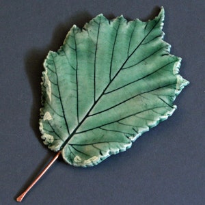 May include: A ceramic leaf sculpture with a green glaze and a copper-colored stem. The leaf has a detailed vein pattern and a slightly textured surface.