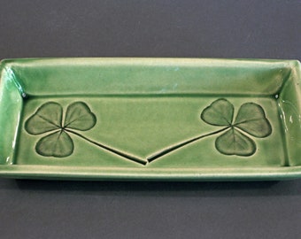 May include: A rectangular, light green ceramic tray with a glossy finish. The tray features two dark green shamrock designs, one on each side, connected by a central stem. The tray has slightly raised edges.