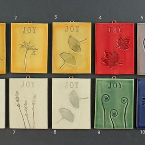 May include: Ten ceramic tiles with the word "JOY" printed on each. Each tile features a different botanical design, including leaves, ferns, and flowers. The tiles are in various colors, including yellow, red, green, blue, and purple.