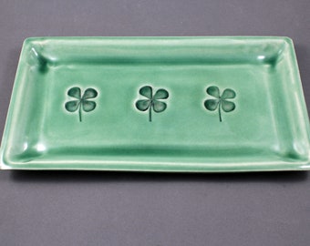 May include: A rectangular, light green ceramic tray with rounded corners. Three embossed shamrock designs are evenly spaced across the tray's surface. The tray has a glossy finish and is set against a gray background.