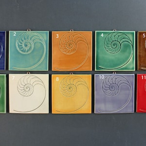 May include: Eleven ceramic tiles with a nautilus shell design in various colors, including blue, green, yellow, brown, and red. The tiles are arranged in two rows of five and one row of one.
