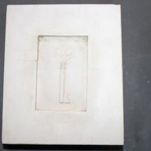 May include: A rectangular, off-white plaster mold featuring a raised relief of a skeleton key. The key design is centered within a recessed rectangular frame, with a detailed bow and a long, slender stem. The mold has a slightly aged appearance.