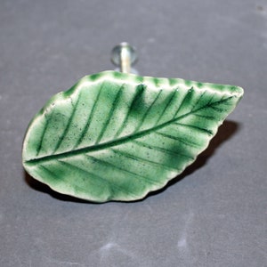 May include: A green ceramic leaf-shaped cabinet knob with a clear glass stem. The leaf has detailed veining and a glossy finish. This decorative hardware adds a natural touch to furniture.
