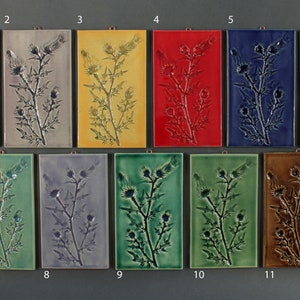 May include: Eleven ceramic tiles with a raised design of a thistle plant. The tiles are arranged in two rows, with the top row featuring tiles in white, gray, yellow, red, blue, and brown. The bottom row features tiles in green, gray, green, green, brown, and brown.