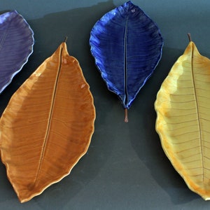 May include: Four ceramic leaf-shaped serving dishes in shades of purple, orange, and yellow. The dishes are glazed and have a realistic leaf texture.
