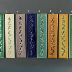 May include: Ten ceramic tiles with a raised design of a single stalk of wheat. The tiles are arranged in a row from left to right, each tile is a different color: red, green, white, purple, blue, teal, orange, yellow, green, and brown.
