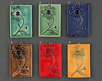 May include: Six ceramic tiles with a raised design of a single flower with a black center and green leaves. The tiles are in six different colors: teal, green, blue, brown, red, and yellow.