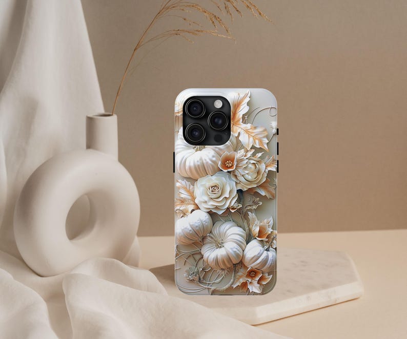 May include: A white iPhone case with a 3D floral design. The case features pumpkins and roses in white and peach tones. The case has a glossy finish and a black camera module.