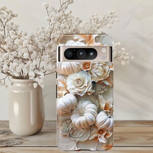 May include: A phone case with a 3D floral design. It features white pumpkins, roses, and leaves in white, cream, and orange hues. The case is on a wooden surface, with a vase of white flowers in the background.