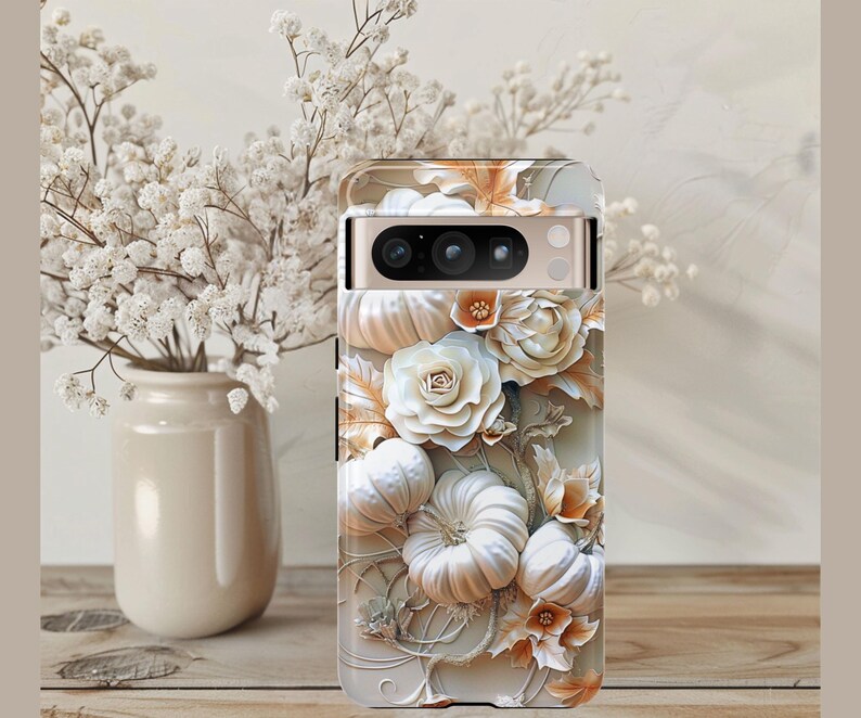 May include: A phone case with a 3D floral design. It features white pumpkins, roses, and leaves in white, cream, and orange hues. The case is on a wooden surface, with a vase of white flowers in the background.