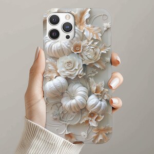 May include: A phone case with a 3D design of white pumpkins, roses, and leaves. The case has a light grey background and a textured appearance. The design includes intricate details and a seasonal autumn theme.