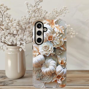 May include: A phone case with a 3D floral design. It features white pumpkins, roses, and leaves in white, cream, and gold. The case has a black camera module. The background is neutral.