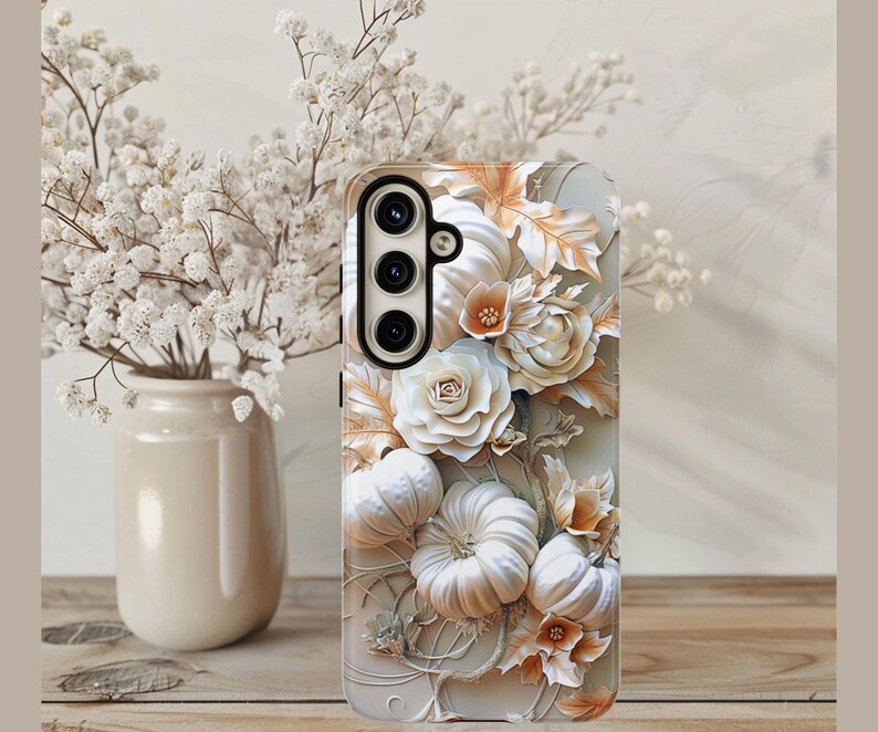 May include: A phone case with a 3D floral design. It features white pumpkins, roses, and leaves in white, cream, and gold. The case has a black camera module. The background is neutral.