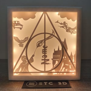 Puede incluir: Caja de sombras cuadrada e iluminada con siluetas recortadas de un coche, un búho, un mago, un castillo y el símbolo de las Reliquias de la Muerte. La caja es blanca con un interior cálido y brillante. El texto "STC 3D" es visible.