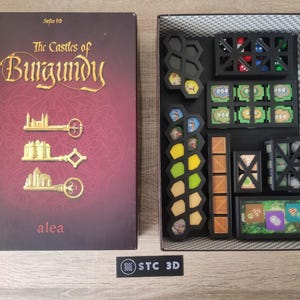 May include: The Castles of Burgundy board game box with the game's title and logo on the front. The box is open and shows the game's components, including wooden pieces, tiles, and cards.