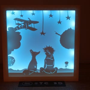 Puede incluir: Un marco blanco iluminado con un fondo azul que representa una silueta del Principito con un zorro, un avión y estrellas. El marco está iluminado desde atrás, creando un efecto brillante.