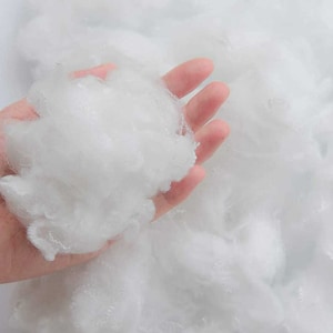 May include: A close-up of a hand holding a large amount of white, fluffy fibrefill. The fibrefill is soft and appears to be made of polyester.
