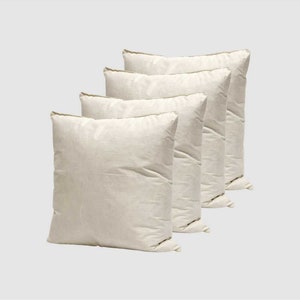 May include: Four off-white square throw cushions with a subtle texture. The cushions are arranged in a row, with the largest cushion at the back and the smallest cushion at the front.